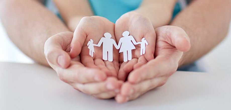 Das Bild zeigt vier Hände: Unten liegen die größeren Hände, vermutlich eines Mannes, nebeneinander mit den Handinnenflächen nach oben. Darüber sind ebenso zwei Hände nebeneinander gelegt. In den oberen Handinnenflächen befindet sich ein Papierzuschnitt, der eine Familie darstellen soll.