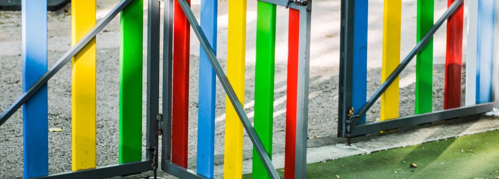 Das Bild zeigt ein offenes, buntes Gartentürchen, welches von einem Schotteruntergrund zu einem grünen Untergrund führt.