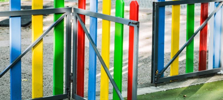 Das Bild zeigt ein offenes, buntes Gartentürchen, welches von einem Schotteruntergrund zu einem grünen Untergrund führt.