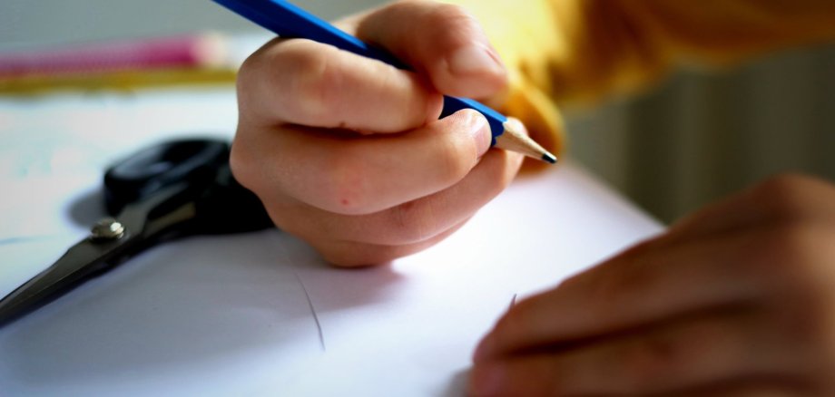 Das Bild zeigt die Hand eines Kindes, die einen Bleistift hält. Darunter befindet sich ein weißes Blatt Papier.