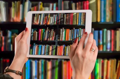 Auf dem Bild ist ein Bücherregal. Vor dem Bücherregal steht eine Person mit einem Ipad und macht ein Bild von dem Bücherregal.