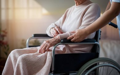 Auf dem Bild ist eine Person zu sehen, die in einem Rollstuhl sitzt. Sie wird von einer anderen Person geschoben.