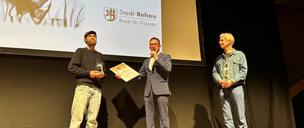 Auf dem Bild sind Bürgermeister Michael Abraham und Thorsten Schaumann neben dem diesjährigen Hans-Vogt-Filmpreisträger Robert Gwisdek zu sehen, der gerade die Urkunde und den Preis von Bürgermeister Abraham auf der Bühne entgegennimmt.