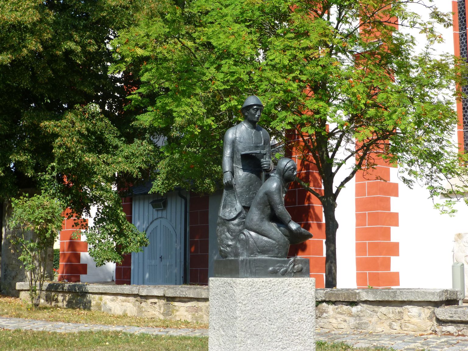 Das Bild zeigt das Kartoffeldenkmal, das vor der Kirche in Pilgramsreuth steht. Darauf sind ein Mann und eine Frau beim Kartoffel graben zu erkennen.