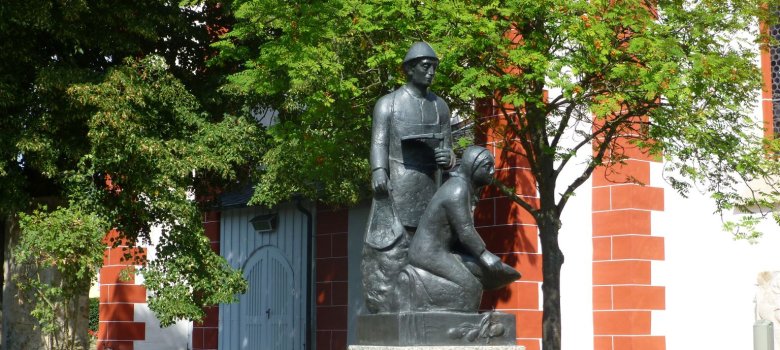 Das Bild zeigt das Kartoffeldenkmal, das vor der Kirche in Pilgramsreuth steht. Darauf sind ein Mann und eine Frau beim Kartoffel graben zu erkennen.