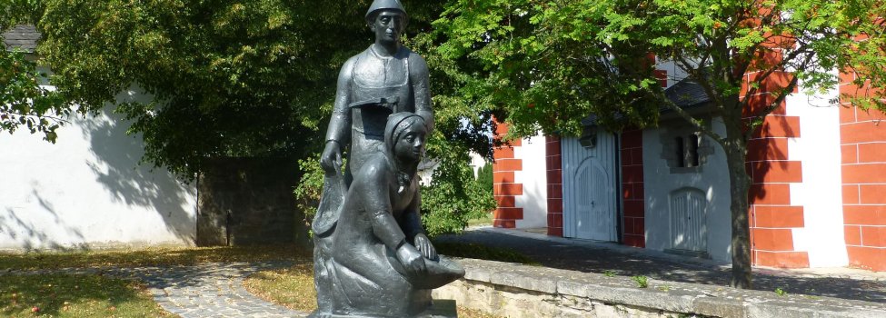 Das Bild zeigt das Kartoffeldenkmal, das vor der Kirche in Pilgramsreuth steht. Darauf sind ein Mann und eine Frau beim Kartoffel graben zu erkennen.