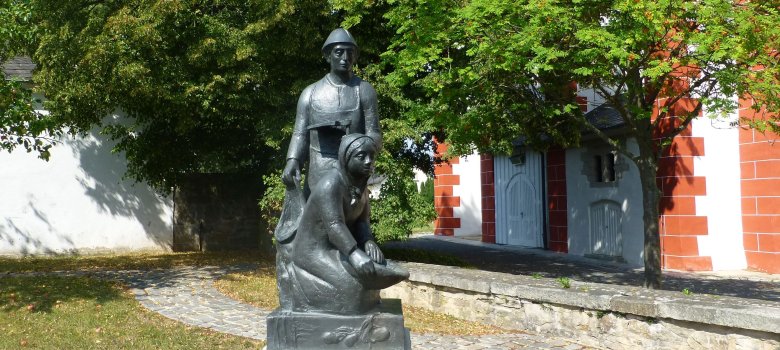 Das Bild zeigt das Kartoffeldenkmal, das vor der Kirche in Pilgramsreuth steht. Darauf sind ein Mann und eine Frau beim Kartoffel graben zu erkennen.