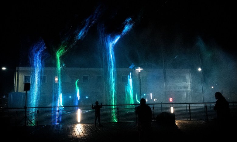 Das Bild zeigt den Blick vom Maxplatz zum Perlenbach bei Nacht. Wasserfontänen am Perlenbach sind grün/blau beleuchtet.
