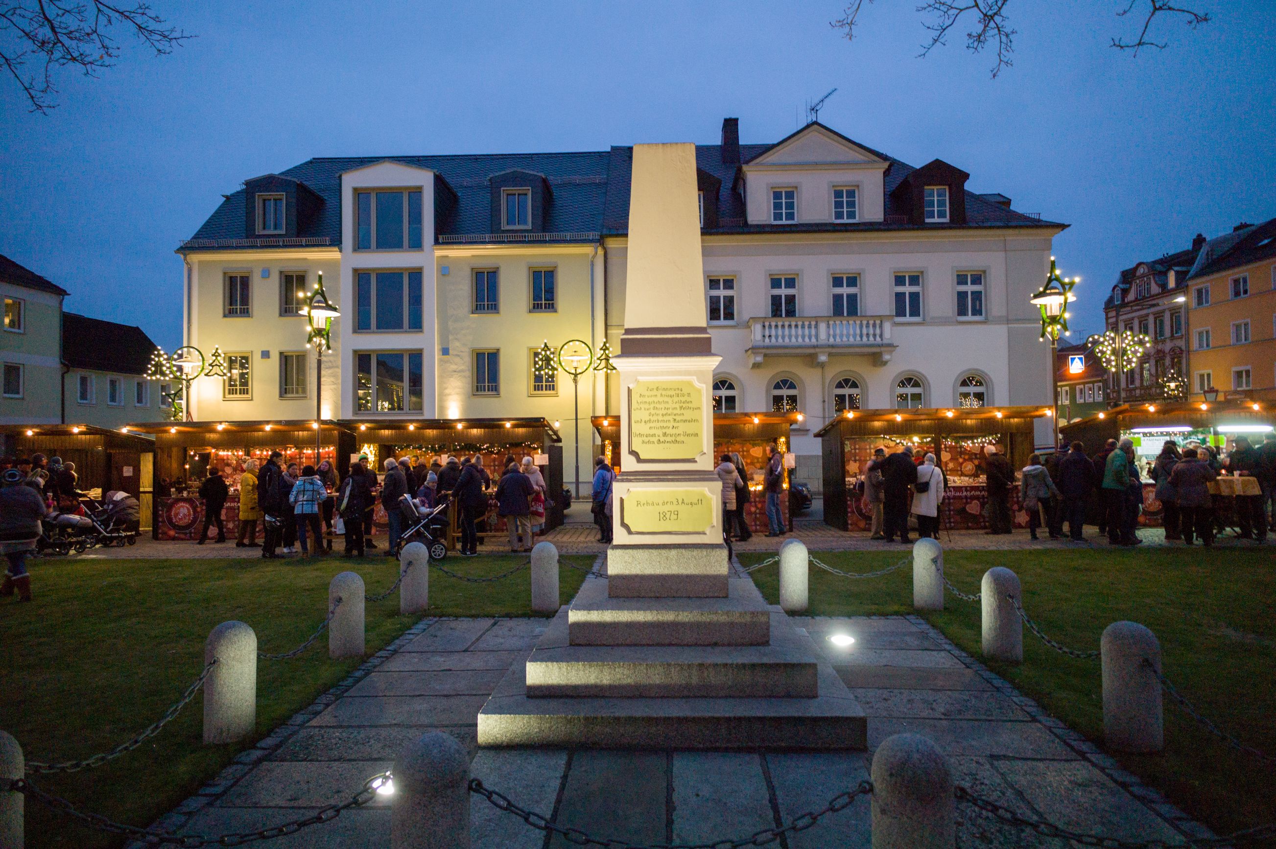 Auf dem Bild ist vordergründig ein Denkmal auf dem Rehauer Maxplatz zu erkennen. Im Hintergrund befinden sich einige Besucher vor Holzhütten auf dem Rehauer Lebkuchenmarkt.