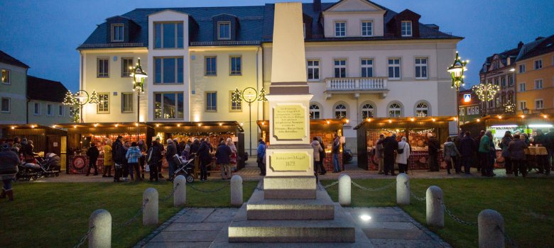 Auf dem Bild ist vordergründig ein Denkmal auf dem Rehauer Maxplatz zu erkennen. Im Hintergrund befinden sich einige Besucher vor Holzhütten auf dem Rehauer Lebkuchenmarkt.