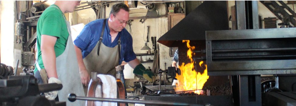 Das Bild zeigt die Mechanischer Werkstatt von innen. Darauf ist die brennende Schmiede zu sehen, welche von einem Handwerker bedient wird.