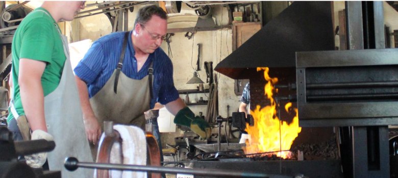 Das Bild zeigt die Mechanischer Werkstatt von innen. Darauf ist die brennende Schmiede zu sehen, welche von einem Handwerker bedient wird.