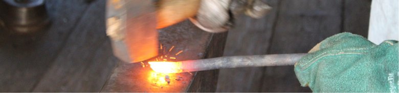Auf dem Bild ist zu sehen, wie jemand schmiedet. Konkret hält eine Hand einen Metallstab fest während ein Hammer darauf klopft.