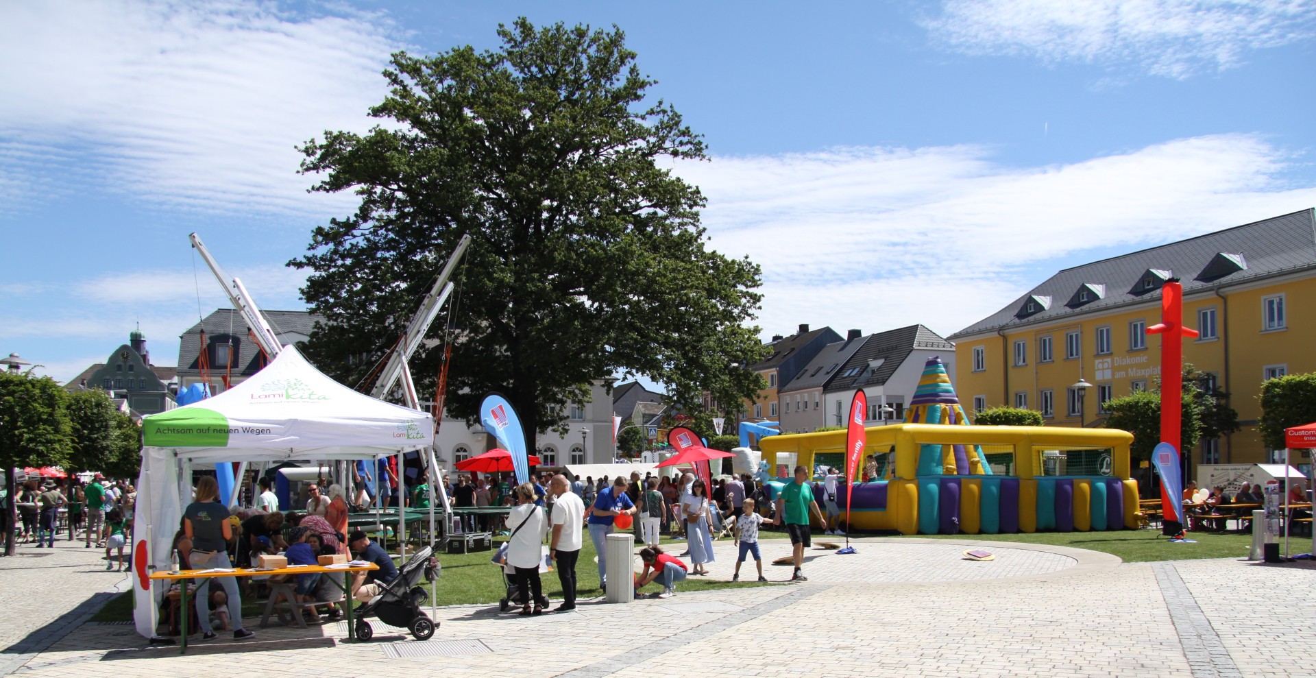 Das Bild zeigt verschiedene Vergnügungsattraktionen, welchen mitten auf dem Maxplatz in Rehau aufgebaut sind. Daneben sind noch Sitzgelegenheiten und Besucher des Rehauer Stadtfests zu erkennen.