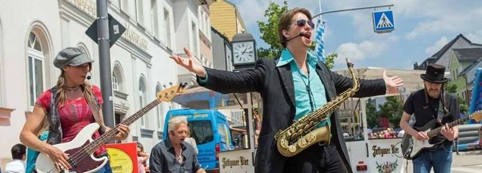 Das Bild zeigt Musiker mitten in Rehau. Konkret handelt es sich um vier Musiker, die auf einer Straße in der Innenstadt von Rehau spielen. Im Hintergrund ist ein Schankwagen sowie Vergnügungsattraktionen zu erkennen.