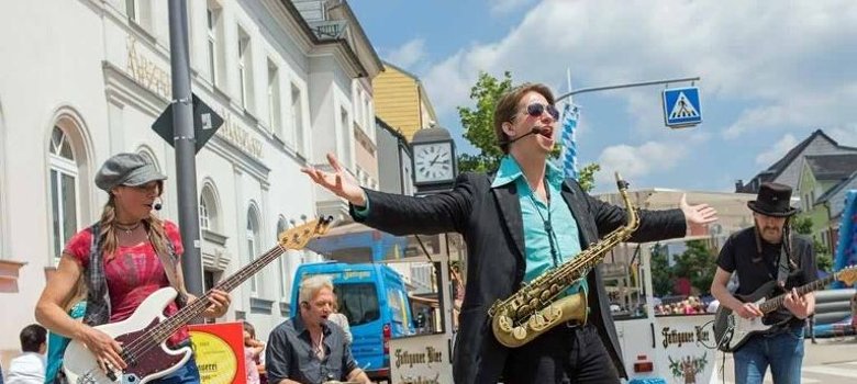 Das Bild zeigt Musiker mitten in Rehau. Konkret handelt es sich um vier Musiker, die auf einer Straße in der Innenstadt von Rehau spielen. Im Hintergrund ist ein Schankwagen sowie Vergnügungsattraktionen zu erkennen.