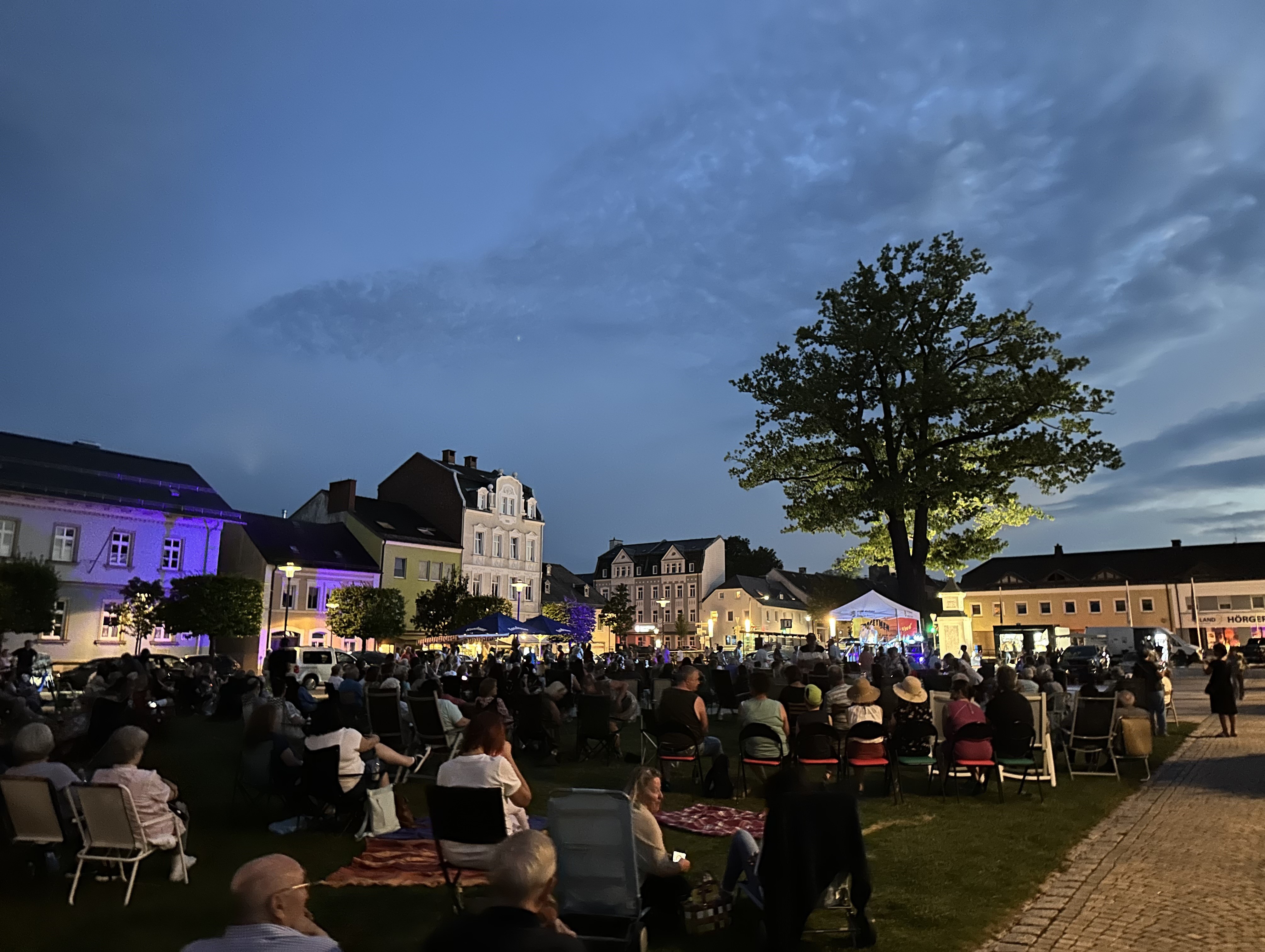 Das Bild zeigt den mit Menschen gefüllten Maxplatz während eines Stuhlkonzerts. Es wurde kurz vor Einbruch der Dunkelheit aufgenommen während eine Band auf der Bühne spielt.