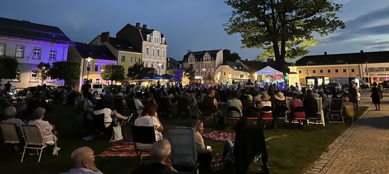 Das Bild zeigt den mit Menschen gefüllten Maxplatz während eines Stuhlkonzerts. Es wurde kurz vor Einbruch der Dunkelheit aufgenommen während eine Band auf der Bühne spielt.