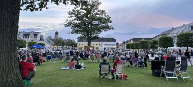 Das Bild zeigt den mit Menschen gefüllten Maxplatz bei einem Rehauer Stuhlkonzert. Die Besucher sitzen auf ihren eigens mitgebrachten Sitzgelegenheiten auf der Wiese. Davor spielt eine Band auf der Stuhlbühne.