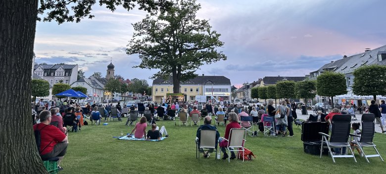 Das Bild zeigt den mit Menschen gefüllten Maxplatz bei einem Rehauer Stuhlkonzert. Die Besucher sitzen auf ihren eigens mitgebrachten Sitzgelegenheiten auf der Wiese. Davor spielt eine Band auf der Stuhlbühne.