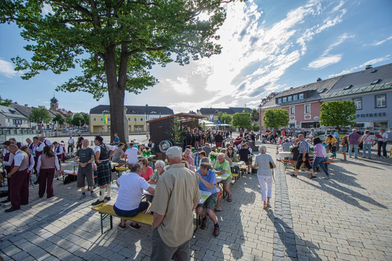 Das Bild zeigt einige Besucher auf Bierzeltgarnituren sitzend auf dem Maxplatz. Links auf dem Bild sind Musiker des Rehauer Musikvereins zu erkennen.