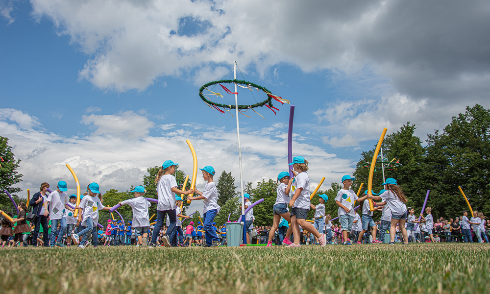 Das Bild zeigt Kinder bei den traditionellen Aufführungen am Rehauer Wiesenfest nach dem Umzug. Die Kinder haben weiße T-Shirts und blaue Hosen an. Dahinter sind zahlreiche Zuschauer zu sehen.