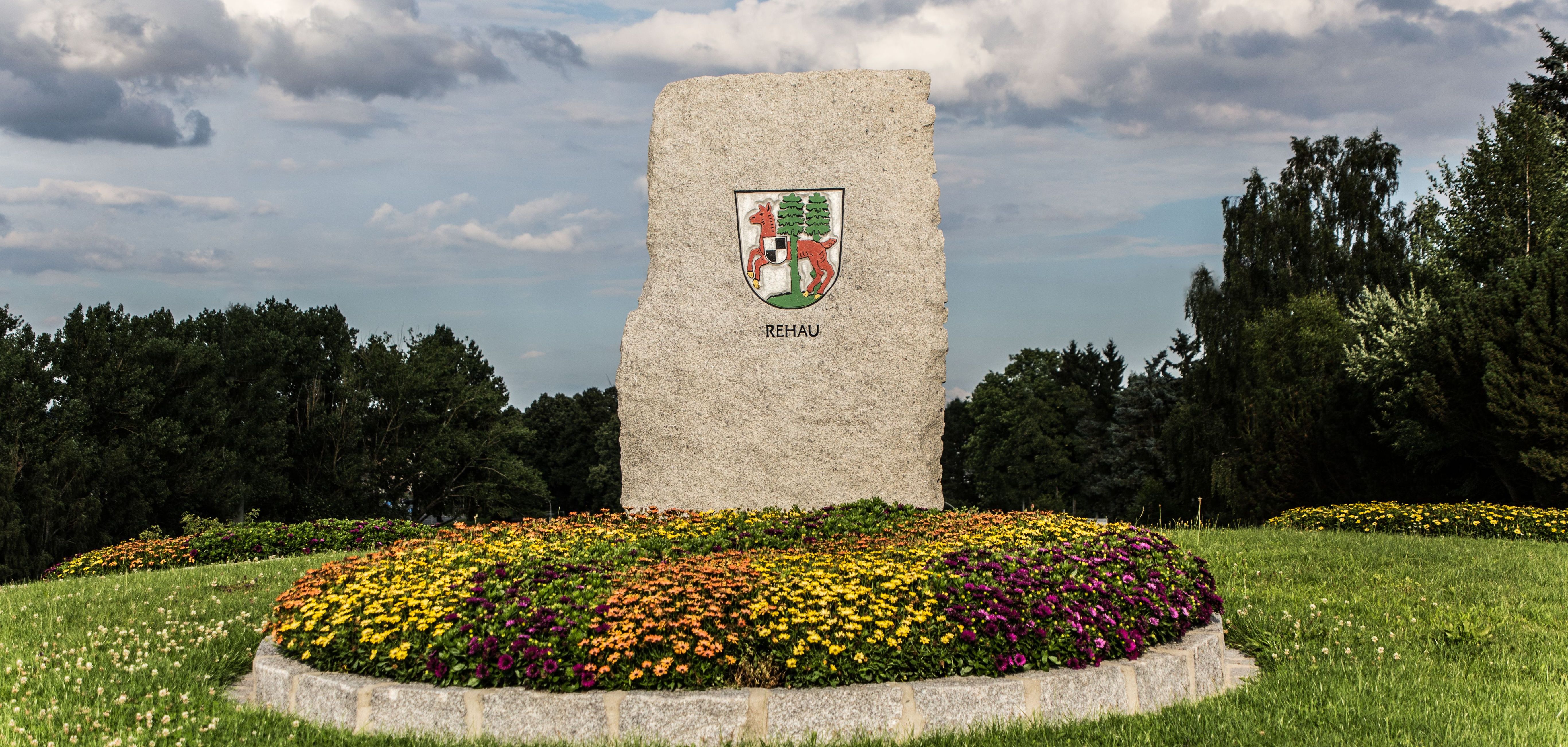 Das Bild zeigt den Kreisverkehr am Ortseingang von Rehau. Darauf befindet sich ein großer Stein mit dem Wappen der Stadt Rehau, davor ein schön angelegtes, buntes Blumenbeet.