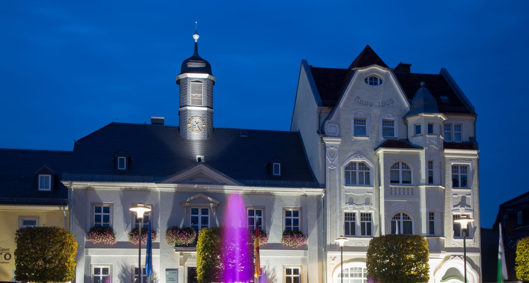 Das Bild zeigt die Anwesen Maxplatz 5 und 7 bei Nacht. Davor ist noch eine Fontäne des beleuchteten Brunnens zu erkennen.