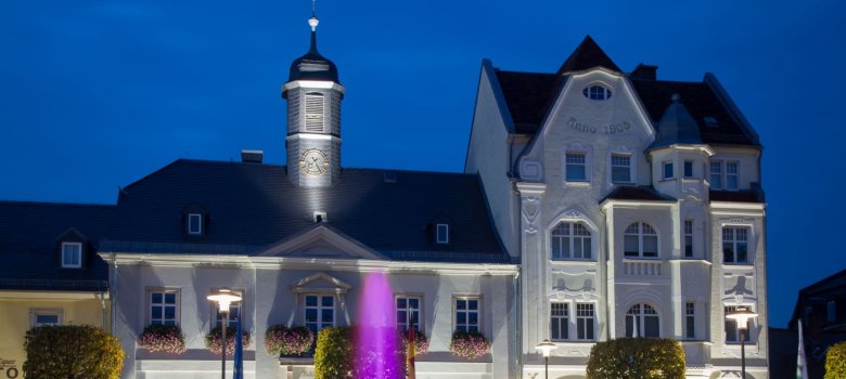 Das Bild zeigt die Anwesen Maxplatz 5 und 7 bei Nacht. Davor ist noch eine Fontäne des beleuchteten Brunnens zu erkennen.