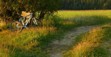 Das Bild zeigt eine Kurve in einem Feldweg. Links davon befindet sich eine Holzbank und ein davor abgestelltes Fahrrad. Die Lichtverhältnisse lassen einen kurz bevorstehenden Sonnenuntergang vermuten.