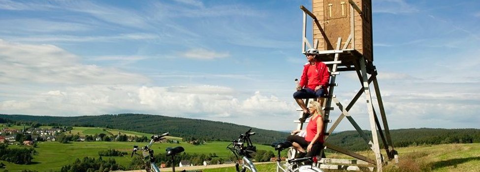 Das Bild zeigt zwei Fahrradfahrer, die gerade auf einem Jägerhochstand eine Pause einlegen. Die Fahrräder haben Sie vor abgestellt. Im Hintergrund sind Wiesen, Felder, Wälder und ein paar Häuser zu erkennen.