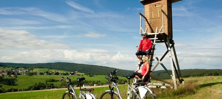 Das Bild zeigt zwei Fahrradfahrer, die gerade auf einem Jägerhochstand eine Pause einlegen. Die Fahrräder haben Sie vor abgestellt. Im Hintergrund sind Wiesen, Felder, Wälder und ein paar Häuser zu erkennen.
