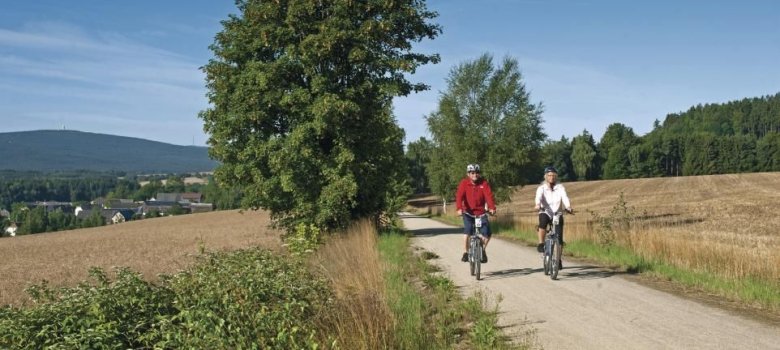 Auf dem Bild sind zwei Fahrradfahrer auf einem schönen Fahrradweg zwischen Felden zu sehen. Im Hintergrund ist ein Berg sowie einige Häuser zu erkennen.
