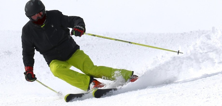 Das Bild zeigt einen Skifahrer mit grüner Hose, schwarzer Jacke und einem Helm auf einer Skipiste bei einem Schwung.
