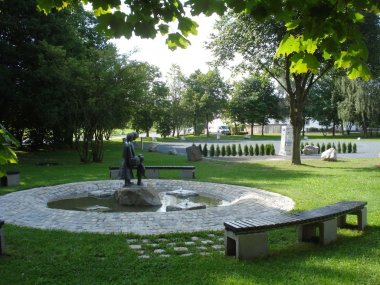 Auf dem Bild ist der Brunnen am Siedlerfestplatz zu sehen. Um den Brunnen befinden sich ein paar Bänke. Im Hintergrund ist der neu angelegte, geschotterte Wohnmobilpark mit dem Automaten sowie neu angepflanzten Bäumen zu erkennen.