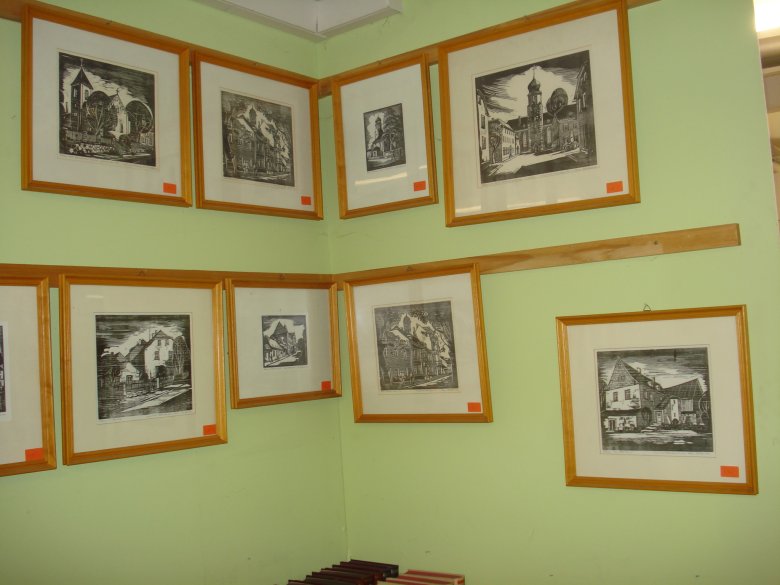 Das Bild zeigt verschiedene Zeichnungen in Holzrahmen die an der grünen Wand befestigt sind.