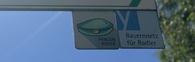 Auf dem Bild ist ein Fahrradschild zu sehen, unter welchem ein kleines Schild mit dem Logo der Perlenroute ist. 